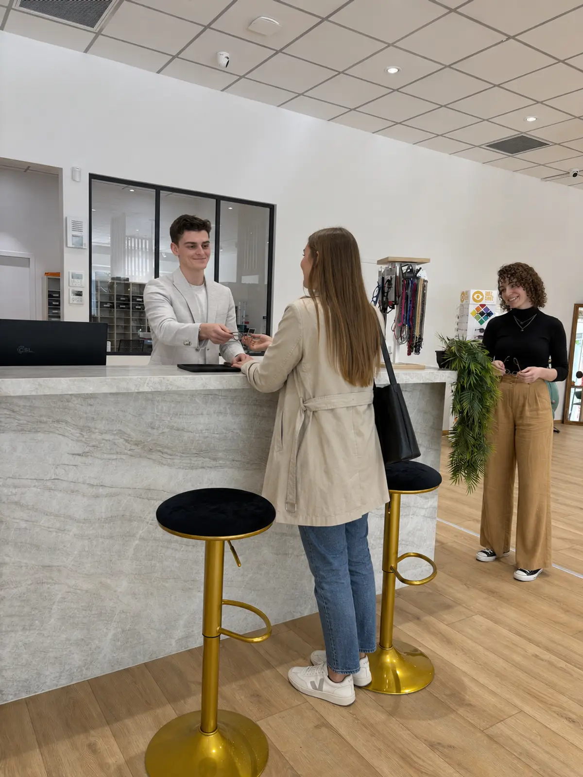 Au comptoir, Nathan accueille une cliente ; Chloé prépare une monture.