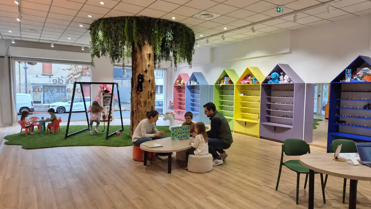Une famille choisit des lunettes sous l'arbre géant de l'espace enfant, devant les maisons colorées.